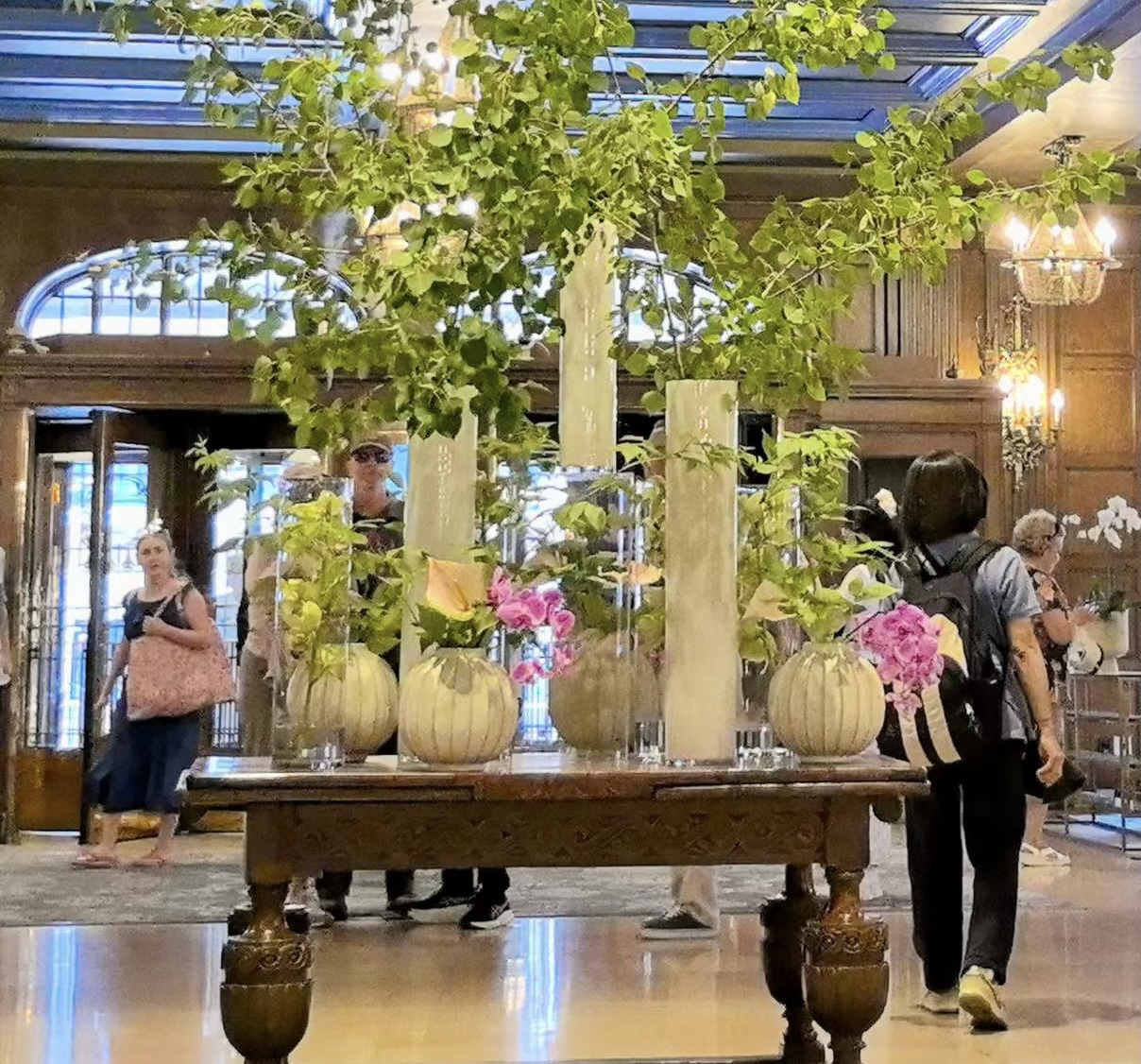 A floral arrangement of green branches and pink flowers in tall vases on a carved wooden table in a hotel lobby.